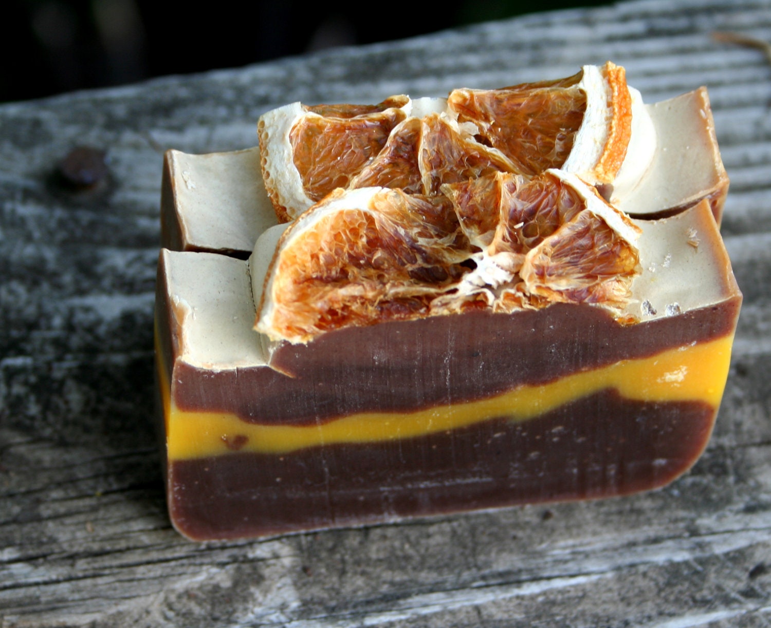 Two bars of soap that look like slices of chocolate orange cake: a half-orange slice on top of a white frosting-looking layer on top of an orange layer between two dark chocolate-colored layers.
