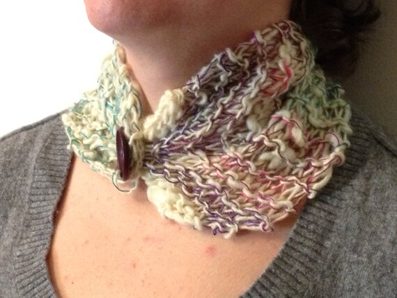 Handspun/hand-knit cowl with cream as the primary color and added accents of blue/teal, purple and pink/magenta. Worn by a model who shows only the neck/shoulders; the model is wearing a grey sweater and standing in front of a white background.