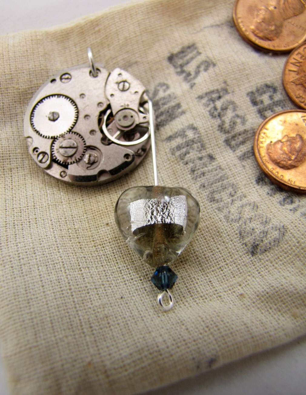pendant made of dismantled clockwork with a pin dangling from it holding a clear and foil glass heart, with deep blue swarovski crystal accent beneath the heart