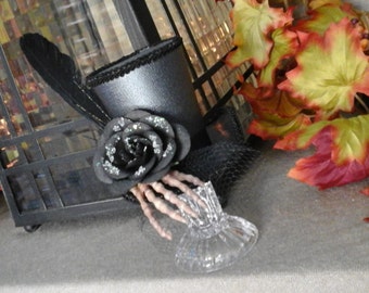 Black mini top hat with skeleton hand and black glitter flower, this piece is attached to a glass stand