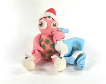 pink 3-eyed polymer clay monster wearing bunny slippers & cuddling a blue blanket