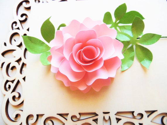 A delicate light pink paper Diana Rose site amid it's paper leaves on top of an ornate wood frame. 