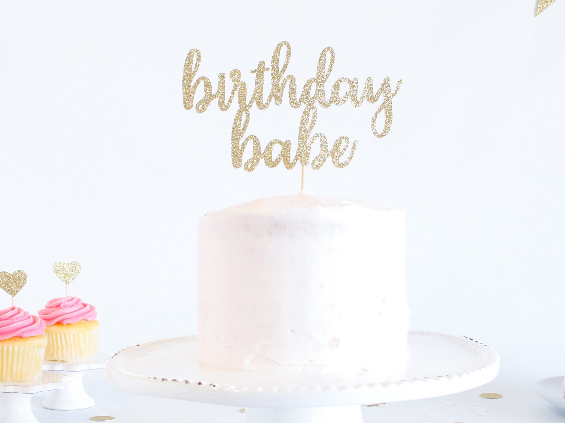 Gold glitter birthday cake topper in top of a white cake in front of a white background. Two pink cupcakes with gold heart toppers are on the left. 